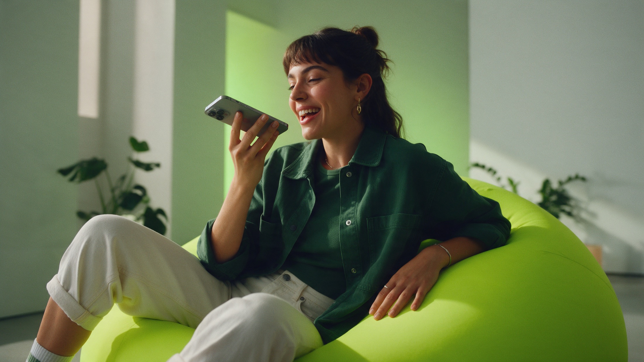 Mulher jovem sentada em um pufe verde, sorridente, conversando com o celular